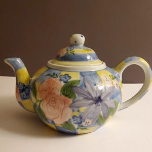 Puede incluir: Una tetera de cerámica con tapa, boquilla y asa. La tetera está pintada con un diseño floral con rosas rosas y flores azules sobre un fondo a cuadros amarillo y azul. El asa y la boquilla son amarillas y azules.