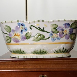Peut inclure: Jardinière en céramique crème avec un bord festonné, ornée de pensées peintes à la main en violet, bleu et jaune et de feuilles vertes. La jardinière a une bande jaune et une base avec des détails d'herbe verte. Décoration.