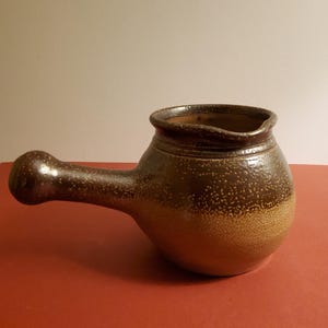 Könnte beinhalten: Handgefertigter, brauner Keramikkrug mit rundem Körper und langem, gebogenem Griff. Der Krug hat eine gesprenkelte Textur und eine weite Öffnung. Der Farbverlauf geht von Dunkelbraun zu einem helleren Beige am Boden über.