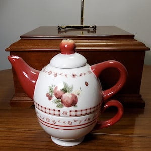 Könnte beinhalten: Eine weiße Keramik-Teekanne mit rotem Griff, Ausguss und Deckel. Die Teekanne hat ein Design aus roten Äpfeln und grünen Blättern. Eine passende Teetasse ist am Boden befestigt. Die Teekanne steht auf einer Holzoberfläche.