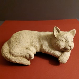 May include: A white ceramic sculpture of a sleeping cat. The cat is curled up on its side with its head resting on its paws. The sculpture is detailed with fur and facial features.