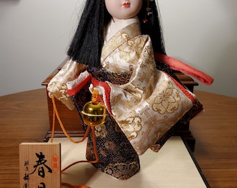 May include: A Japanese doll with straight black hair and a pale face, wearing a kimono with gold and white floral patterns over a dark purple base. The doll holds a gold bell on a cord and stands next to a wooden plaque with black Japanese text.