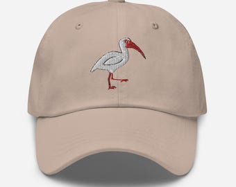 Pode incluir: Um boné de beisebol bege com um ibis branco bordado com bico e pernas vermelhas.