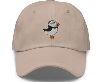 May include: A beige baseball cap with an embroidered image of a puffin. The puffin is white with a black head and beak, and orange feet.