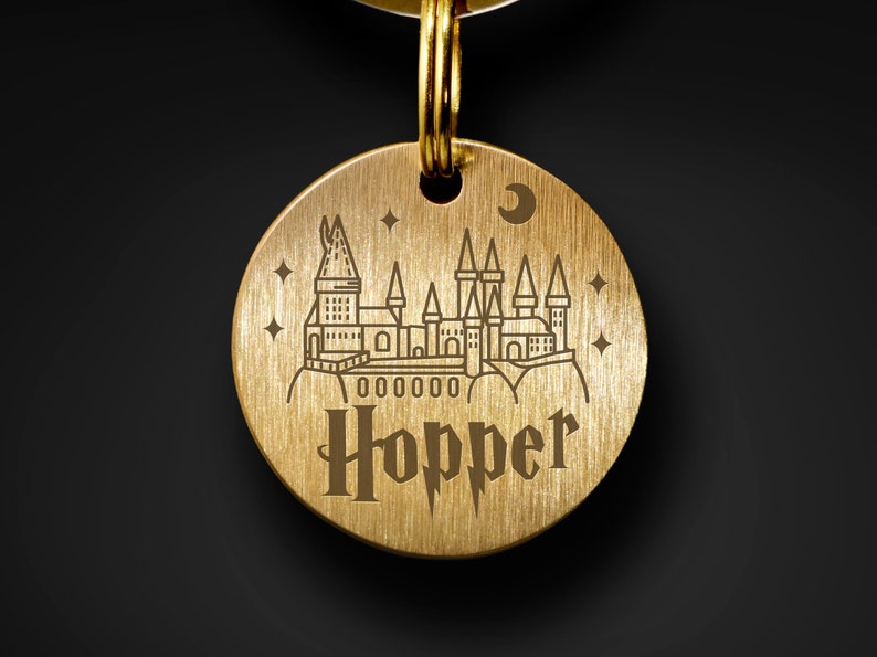 May include: A gold-coloured metal dog tag featuring a Hogwarts castle graphic and the name "Hopper" engraved on it.