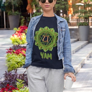May include: A person wearing a black t-shirt with a green and gold chakra symbol and the word "YAM" printed on it. They are wearing a denim jacket and grey sweatpants.