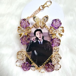 May include: A decorative photo holder featuring a picture of a person. The holder is adorned with gold-colored butterfly and bow accents, along with purple and gold rose embellishments. A gold chain and clasp are attached for hanging.