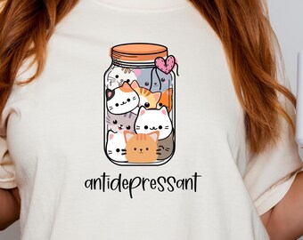 May include: A white t-shirt with a cartoon image of a glass jar filled with cute cartoon cats. The text "antidepressant" is printed below the jar.
