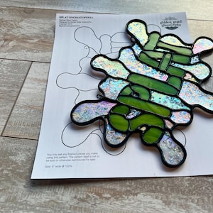 Peut inclure: Une œuvre d'art en vitrail en forme de tache verte avec des accents irisés. La pièce mesure 23 cm de large et est accompagnée d'un modèle pour le design. Le modèle comprend le texte "SPLAT ONOΜΑΤΟΟΕΙΑ" et "golden state glassworks".