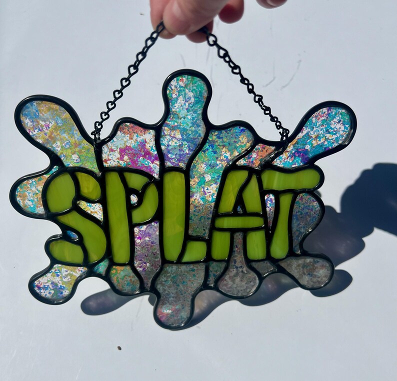 Puede incluir: Una ventana de vitral colgada de una cadena. La ventana tiene forma de salpicadura verde con la palabra "SPLAT" en el centro. El vidrio tiene un acabado iridiscente.