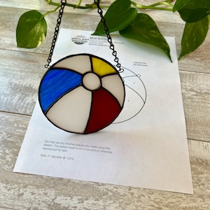Puede incluir: Un atrapasol de vidrio coloreado en forma de pelota de playa con piezas de vidrio azul, rojo, amarillo y blanco. El atrapasol tiene un diámetro de 12,7 cm y cuelga de una cadena negra.