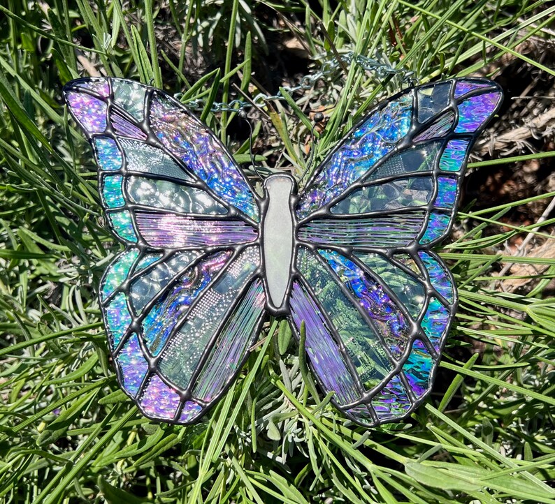 May include: A stained glass butterfly with iridescent purple, blue, and green colors. The butterfly is hanging from a chain and has a white center.