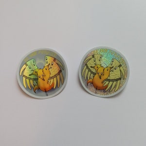 May include: Two round stickers featuring a cartoon illustration of a group of yellow and orange creatures with wings. The stickers have the text "BE NOT AFRAID" around the edge. One sticker has a holographic effect.