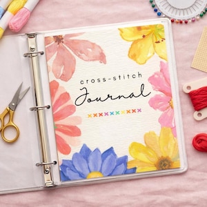 May include: A white binder with a floral cover that reads "cross-stitch Journal." The cover features watercolor flowers in pink, yellow, and blue. Gold scissors and embroidery floss are visible.