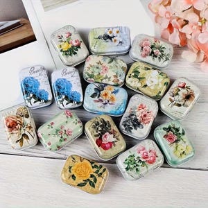May include: A collection of small, rectangular metal tins with floral designs. The tins feature various flowers in shades of pink, yellow, blue, and white against pastel backgrounds. Some tins have the word "Beauty" printed on them.