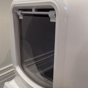 May include: A white, modern cat litter box with a curved design. The front features a clear, rectangular door with a handle. The interior is dark, and the overall aesthetic is clean and minimalist.
