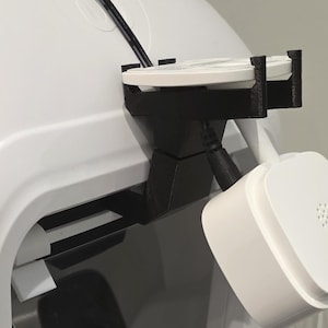 May include: A black 3D-printed mounting bracket attached to a white device. The bracket holds a circular white component and features a black power cable connected to a square white sensor unit with a perforated speaker grille on its front face.