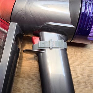 May include: Close-up of a cordless vacuum cleaner. The vacuum features a silver body with red and purple accents. A gray 3D-printed clip is attached to the handle. The image is taken on a wooden surface.