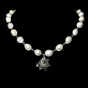 May include: A white pearl necklace with a silver chain and a seashell pendant. The pendant is a detailed, spiky design with black and white patterns. The pearls are irregularly shaped and strung together, creating a classic and elegant look.
