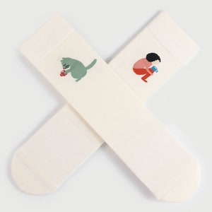 May include: A pair of white socks with embroidered illustrations. One sock features a green cat holding a red flower. The other sock features a person in red and blue clothing holding a blue cup.