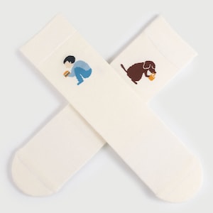 May include: A pair of white socks with embroidered illustrations. One sock features a person sitting and eating a burger, while the other sock features a dog sitting and drinking from a cup.