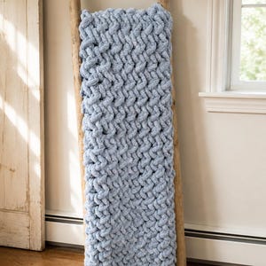 May include: A light blue, chunky knit blanket draped over a wooden ladder. The blanket has a thick, textured weave, and the ladder is made of natural wood. The scene is set indoors, with a window and a door visible in the background.