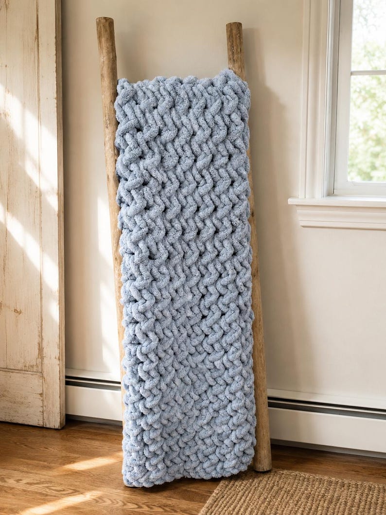 May include: A light blue, chunky knit blanket draped over a wooden ladder. The blanket has a thick, textured weave, and the ladder is made of natural wood. The scene is set indoors, with a window and a door visible in the background.