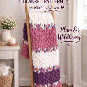 May include: A hand-knitted blanket in shades of plum, white, and wildberry is displayed over a wooden ladder. The blanket pattern is by Wistfully Woven. The chunky knit design features a textured appearance, creating a cosy and inviting aesthetic.