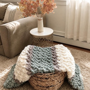 May include: A chunky knit blanket in a woven basket. The blanket features sections of white, gray, and teal. The basket is made of woven wicker and sits on a beige rug. A sofa and side table with a vase of flowers are in the background.