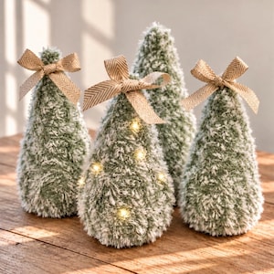 May include: Four cone-shaped decorative trees made of fuzzy, sage green and white textured material. Three of the trees are topped with a tan herringbone patterned ribbon bow. One tree in the front center features small, warm white LED fairy lights.