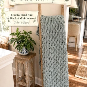 Op de afbeelding: Een dikke, handgebreide deken in een zachte, gedempte blauwgroene kleur, gedrapeerd over een houten ladder. De deken heeft een kruis-herringbone patroon. De afbeelding bevat ook de tekst "Wistfully Woven's Blanket Boot Camp #1: Criss Cross Herringbone" en "Chunky Hand Knit Blanket Mini Course Video Tutorial."