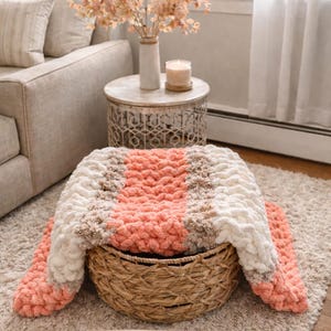 May include: A hand-knitted blanket in a basket, with peach, cream, and tan sections. The image includes the text "Peach Cobbler and Cream" and "Chunky Hand Knit Blanket Pattern by Wistfully Woven • Beginner Friendly". The blanket is a cosy home decor item.