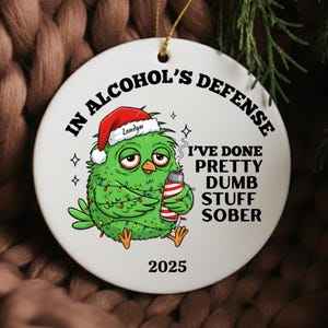 May include: A white ceramic Christmas ornament with the text "IN ALCOHOL'S DEFENSE" and "I'VE DONE PRETTY DUMB STUFF SOBER". It features a green cartoon bird wearing a Santa hat and holding a red and white striped can. The year 2025 is printed at the bottom.