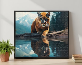 Könnte beinhalten: Ein gerahmtes Puzzle mit einem Puma an einem See mit Bergkulisse. Der Puma ist orange und braun mit grünen Augen, die sich im Wasser spiegeln. Das Puzzle befindet sich in einem schwarzen Rahmen, mit einem Wald und Bergen im Hintergrund.