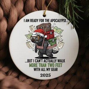 May include: White ceramic Christmas ornament with a cartoon badger wearing a Santa hat and goggles, carrying luggage. Text reads "I AM READY FOR THE APOCALYPSE...BUT I CAN'T ACTUALLY WALK MORE THAN TWO FEET WITH ALL MY GEAR 2025".