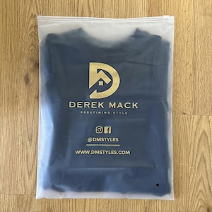 May include: A navy blue item of clothing is sealed in a clear plastic bag. The bag displays gold text and a logo reading "DEREK MACK, REDEFINING STYLE". Social media details and a website address are also printed.