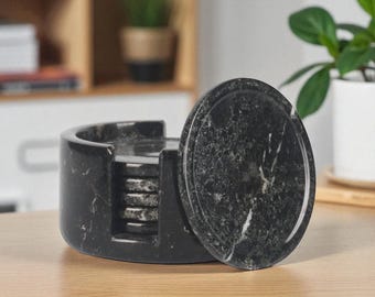 May include: A set of black marble coasters with a matching holder. The coasters are round with a polished finish and white veining. The holder is cylindrical with a curved opening. Home decor.
