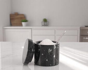 May include: A black marble salt cellar with a matching lid, partially open to reveal a mound of white salt. A small silver spoon rests inside. The container is cylindrical and sits on a white countertop.