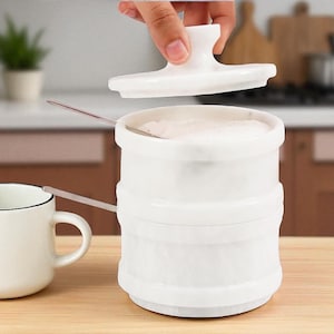 Puede incluir: Un azucarero de cerámica blanca con una tapa que se levanta. El cuenco tiene forma cilíndrica con un diseño acanalado. Una cucharita descansa en el interior. Una taza blanca está en primer plano.