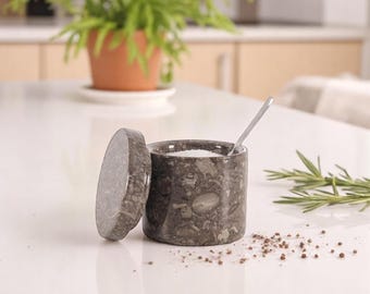 May include: A dark gray marble salt cellar with a matching lid, partially open. A small silver spoon rests inside the container, which is filled with white salt. Pepper and rosemary sprigs are scattered around.