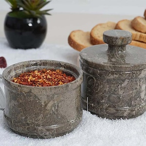 May include: Two gray marble spice jars. One jar is open and filled with red pepper flakes. The other jar has a lid with a round handle. The jars are on a white surface, with a small plant and bread slices in the background.