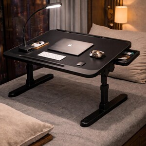 May include: Black adjustable laptop desk with a built-in drawer, a small lamp, and a laptop. The desk is on a bed with pillows. A wireless keyboard and mouse are on the desk.