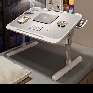 May include: A white and grey adjustable laptop desk with a built-in drawer, a small lamp, and a laptop. The desk also has a keyboard, mouse, and headphones. The desk is designed for comfort and productivity.