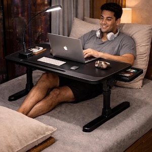 May include: A black adjustable laptop desk with a built-in drawer and a small LED lamp. The desk is set up on a bed, with a laptop, keyboard, and mouse. The person is wearing headphones and shorts.