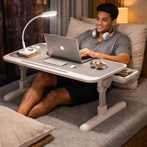 May include: A grey and white adjustable laptop desk with a built-in drawer, a white desk lamp, and a laptop. The desk is supported by white legs and is positioned over a bed. A person is using the laptop.