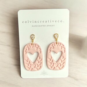 May include: A pair of pink heart-shaped earrings with a gold leaf design and a clear crystal accent. The earrings are handcrafted and are made by colvincreativeco.