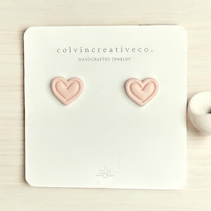 May include: A pair of pink heart-shaped earrings on a white card with the text "colvincreativeco. HANDCRAFTED JEWELRY".