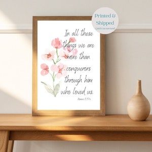May include: A watercolor print with pink flowers and a quote from Romans 8:37a: "In all these things we are more than conquerors through him who loved us."