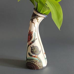 Puede incluir: Un jarrón alto de cerámica blanquecina con cuello curvo, decorado con ilustraciones de búhos marrones y naranjas posados en ramas. El jarrón contiene hojas verdes y flores blancas. El jarrón tiene un borde y una base marrones.