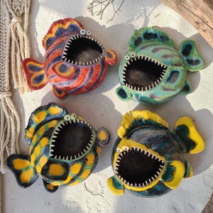 May include: Four ceramic fish-shaped planters with open mouths. Each planter has a unique color scheme: red and blue, green and blue, yellow and blue, and yellow and black. The fish have white teeth and are suitable for small plants or decorative items.